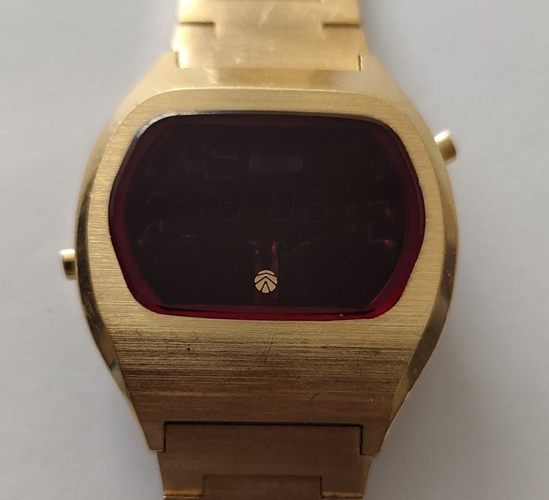 May include: A gold-tone digital wristwatch featuring a brushed metal finish, a rectangular case with rounded edges, and a dark red display screen. The watch face includes a small circular logo at the bottom center and two side buttons on the right.