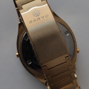 May include: A gold-toned wristwatch featuring a metal link bracelet and a fold-over clasp. The clasp is engraved with the SANYO brand name and a circular logo. The watch case is round with a metallic finish, showing a partial view of the back.