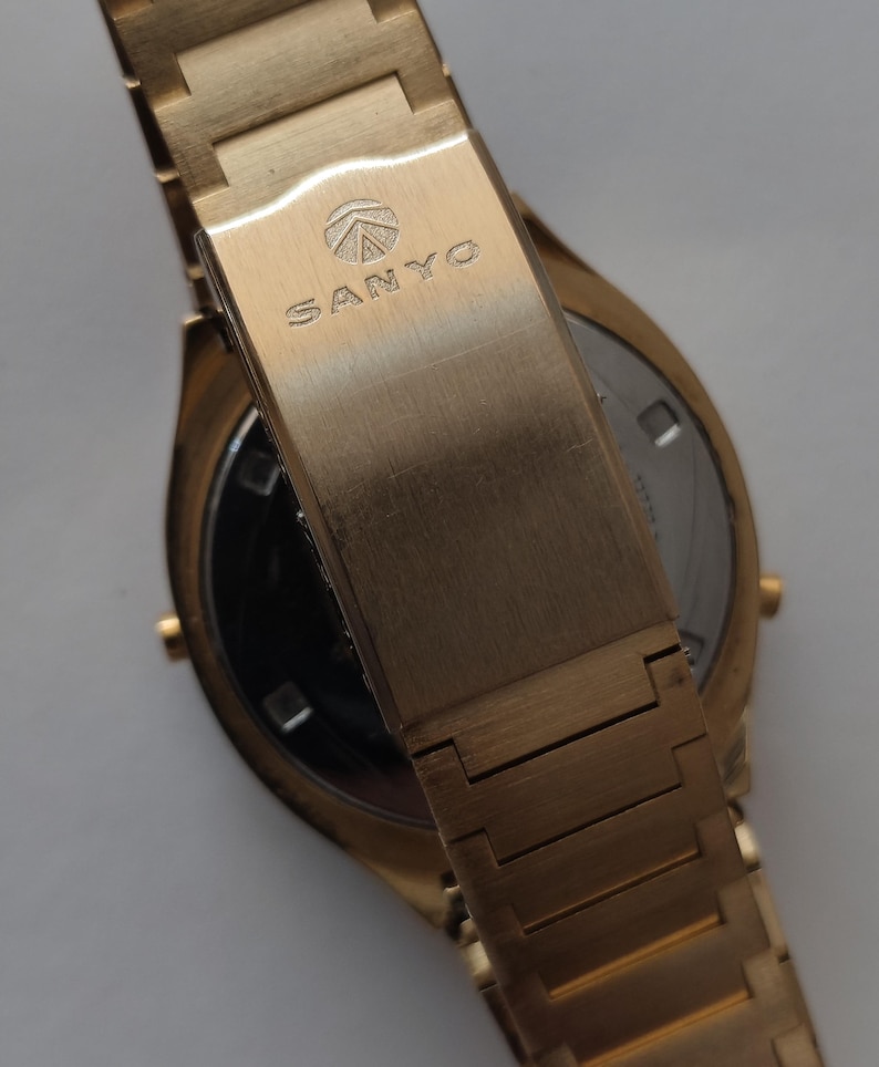 May include: A gold-toned wristwatch featuring a metal link bracelet and a fold-over clasp. The clasp is engraved with the SANYO brand name and a circular logo. The watch case is round with a metallic finish, showing a partial view of the back.