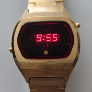 May include: A gold-tone digital wristwatch featuring a rectangular dark red display screen with bright red LED numbers showing 9:55. The watch has a metallic gold-tone band and a small circular logo at the bottom of the display face.
