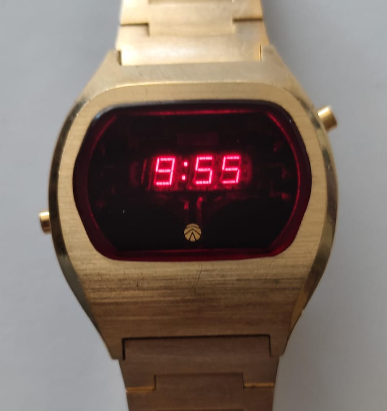 May include: A gold-tone digital wristwatch featuring a rectangular dark red display screen with bright red LED numbers showing 9:55. The watch has a metallic gold-tone band and a small circular logo at the bottom of the display face.