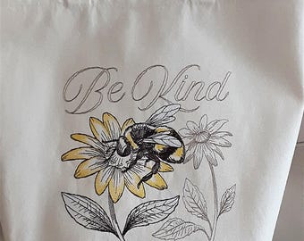 May include: Cream-colored tote bag with two handles. The bag features a detailed illustration of a bumblebee on a yellow and black flower, with the words "Be Kind" in cursive script above the flower. Additional floral elements are also present.