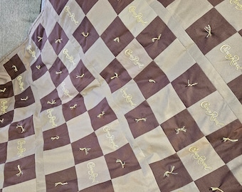 May include: A quilt featuring a checkered pattern of alternating brown and tan fabric squares. Each square is embroidered with the gold text Crown Royal. The quilt is finished with small, light-colored tufted knots at the corners of the squares.