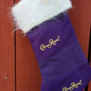 May include: A purple Christmas stocking with the "Crown Royal" logo embroidered in gold. The top of the stocking is white faux fur with a small tan bow. The stocking hangs from a wooden surface.