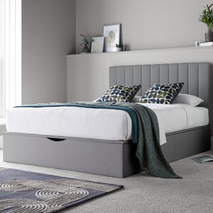 May include: A grey upholstered bed frame with a headboard featuring vertical panels. The bed has a white mattress and a grey storage drawer underneath. A teal blanket is draped over the bed, and two patterned pillows are on top of the mattress.