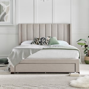 May include: A beige upholstered bed frame with a tufted headboard and a pull-out drawer. The bed is made with a white duvet and a grey throw blanket. There are two decorative pillows on the bed, one with a geometric pattern and one in a solid green colour.