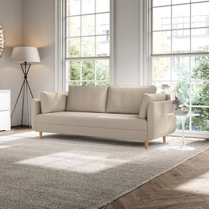 May include: A light beige sofa with wooden legs sits on a light beige rug in a room with large windows. The sofa has two cushions and a light beige fabric upholstery.