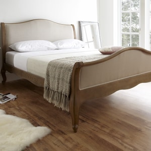 May include: A wooden bed frame with a beige upholstered headboard and footboard. The bed is made of light brown wood and has a curved design. The bed is made up with white sheets, pillows, and a fringed throw blanket. A white rug is on the floor.