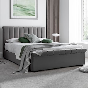 May include: A grey upholstered bed with a vertically panelled headboard and footboard. The bed is made up with white bedding, a grey throw blanket, and decorative pillows in green and patterned grey. A framed landscape painting hangs above the bed.