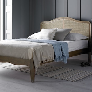 May include: A wooden bed frame with a woven cane headboard. The bed is made up with a white sheet, a light brown blanket, and a blue throw. The bed is sitting on a striped rug.
