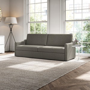 May include: A grey fabric sofa with a tufted back and armrests. The sofa is in a living room with a large rug and windows.