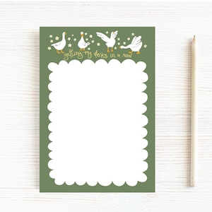May include: A notepad with a green background and a white scalloped border. The top of the notepad features four cartoon ducks and the phrase "getting my ducks in a row" in yellow script. A pencil rests on the right side.