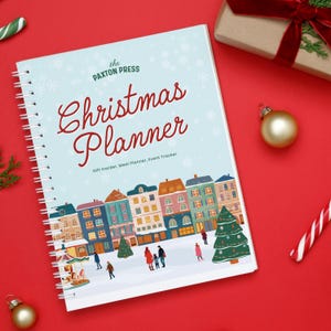 May include: A spiral-bound Christmas Planner with a light blue cover featuring the text "Christmas Planner" in red script. The cover also includes a town illustration, snowflakes, and the text "Gift tracker, Meal Planner, Event Tracker."