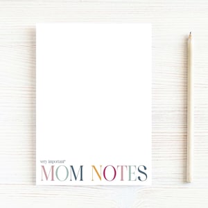 May include: A white notepad with the words "very important" and "MOM NOTES" printed in colorful letters. A wooden pencil rests on the right side of the notepad. The notepad is designed for taking notes.