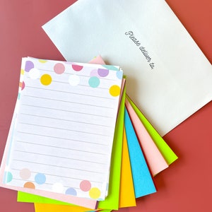 May include: A stack of colorful stationery paper with a polka dot pattern and lined writing paper. The top sheet has a white background with colorful polka dots. The paper is folded in half and has a white envelope with the text "Please deliver to:" on the front.