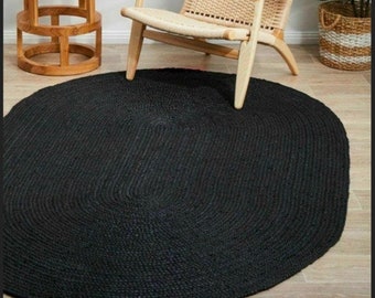 May include: A black oval jute rug with a braided texture. The rug is on a wooden floor and a chair is sitting on top of it.