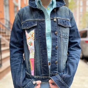 May include: A dark wash denim jacket with a patchwork design on the front. The jacket has a classic collar, button closures, and pockets. The patchwork features floral and geometric patterns in various colors. A light blue and white checkered shirt is visible underneath.