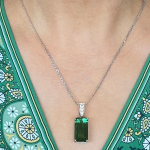 May include: A silver chain necklace with a pendant featuring a large, rectangular, emerald green gemstone surrounded by smaller, clear gemstones.