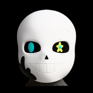 May include: A white skull-shaped figurine with a wide grin and a black, stylized hand on the left side. The left eye has a teal oval, and the right eye features a yellow star. The eye sockets have a mesh texture.