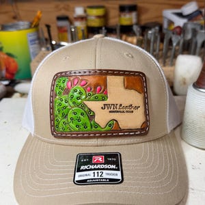 May include: A tan and white trucker hat featuring a rectangular leather patch on the front. The patch displays a green cactus with pink flowers and a tan Texas-shaped cutout with the text JWN Leather Huntsville, Texas. A black Richardson 112 sticker is on the brim.