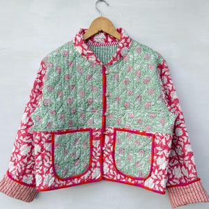 Könnte beinhalten: Eine gesteppte Jacke mit einem hellgrünen Blumenmuster am Oberkörper und an den Taschen, das mit einem leuchtend rosa-weißen Blumenmuster an den Ärmeln und am unteren Teil kontrastiert. Sie hat einen leuchtend rosa Saum, Knopfverschluss und gestreifte Bündchen.