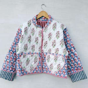 Op de afbeelding: Een gewatteerd jasje met een witte basis en een roze en groene bloemenprint. De mouwen en het onderste gedeelte hebben een blauw en roze bloemenpatroon. Het bevat twee hartvormige voorzakken en een rood-wit gestreepte bies.