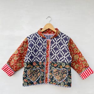 May include: A quilted jacket featuring a mix of patterns including a dark blue and white geometric lattice print on the upper section, a dark floral print on the lower section with two heart-shaped pockets, and red floral sleeves with striped cuffs.
