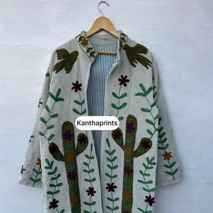 May include: A light-colored, open-front jacket with embroidered cactus and floral designs. The jacket features green, brown, and purple accents, with the text "Kanthaprints" visible. The interior lining has a blue and white striped pattern.