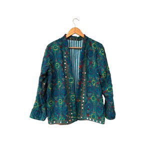 May include: Teal embroidered jacket with floral and mirror details. The jacket is hanging on a wooden hanger against a white background. The jacket has a striped lining and red button closures.