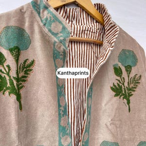 May include: A beige, floral-print jacket with a teal and green floral design. The jacket has a striped brown and white lining and is hanging on a wooden hanger. The text "Kanthaprints" is visible.
