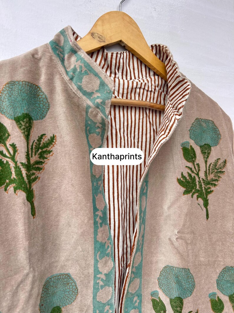 May include: A beige, floral-print jacket with a teal and green floral design. The jacket has a striped brown and white lining and is hanging on a wooden hanger. The text "Kanthaprints" is visible.