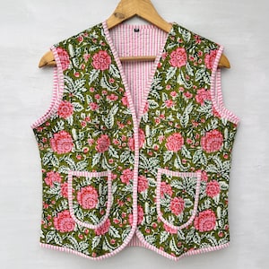 May include: A sleeveless vest with a floral pattern in shades of green and pink. The vest has two front pockets and a pink and white striped lining. The edges are trimmed with pink piping. The vest is hanging on a wooden hanger.
