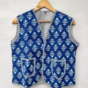 May include: A sleeveless blue vest with a floral pattern and two pockets. The vest has a striped lining and trim. The floral pattern is white and light blue. The vest is hanging on a wooden hanger.