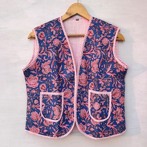 May include: A quilted vest with a blue floral pattern and pink trim. The vest has two front pockets and a pink and white striped lining. It is hanging on a wooden hanger.