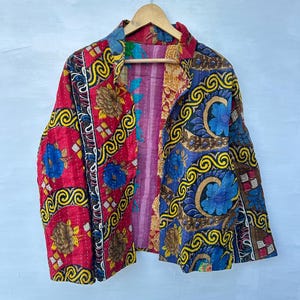 May include: A colorful, quilted jacket with a floral and scroll pattern. The jacket features a mix of red, blue, and yellow hues, with a contrasting pink lining. It is hanging on a wooden hanger against a light background.