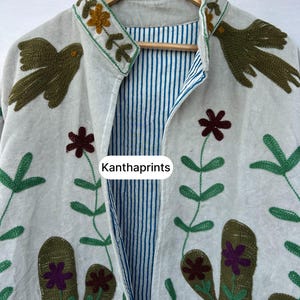 May include: A light-colored jacket with embroidered floral and bird designs. The jacket features a striped blue and white lining and the text "Kanthaprints". The embroidery includes green, brown, and purple accents.