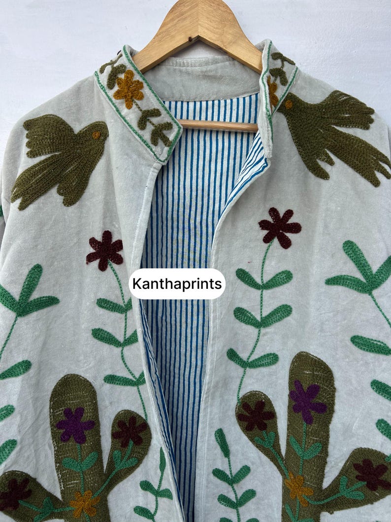 May include: A light-colored jacket with embroidered floral and bird designs. The jacket features a striped blue and white lining and the text "Kanthaprints". The embroidery includes green, brown, and purple accents.