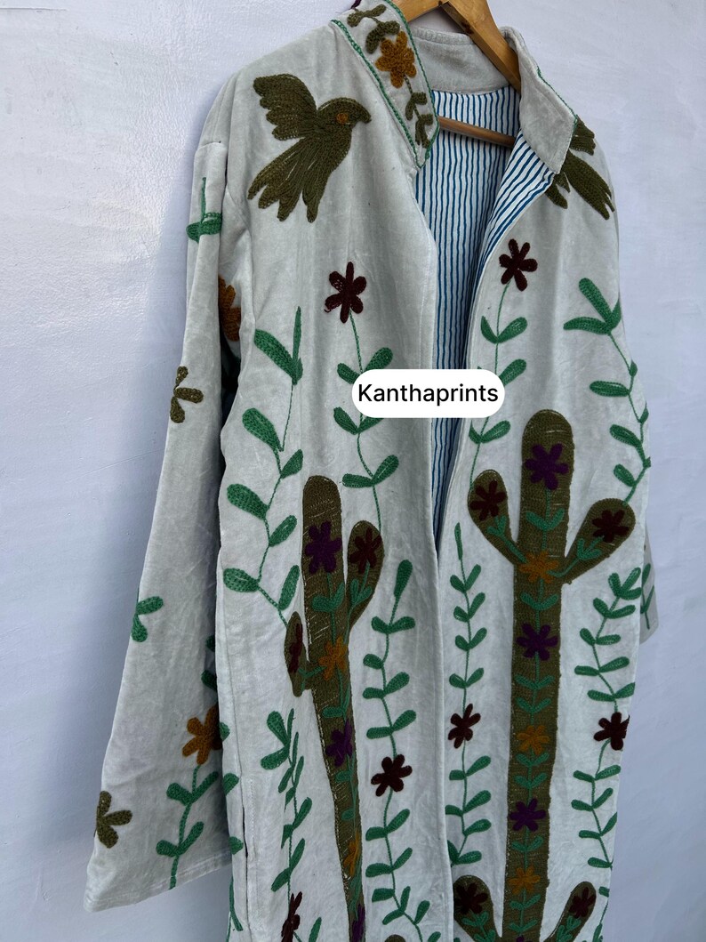 May include: A light gray embroidered jacket with a botanical design. The jacket features a cactus, flowers, and bird embroidery in green, brown, and purple. The inside lining has blue and white stripes. The text "Kanthaprints" is visible.