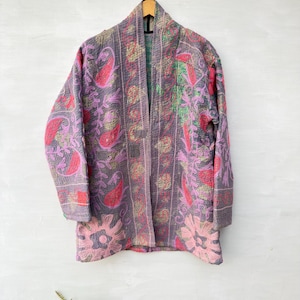 Pode incluir: Um casaco de manga comprida com base cinzenta e bordados intrincados e coloridos de flores e motivos paisley em tons de rosa, verde e amarelo. A peça tem a parte da frente aberta, gola xale e costuras acolchoadas visíveis.