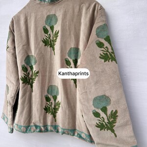 May include: Beige jacket with a high collar and long sleeves, adorned with a repeating pattern of teal floral designs and green leaves. The trim and collar feature a teal and white pattern. The text "Kanthaprints" is visible on the jacket.