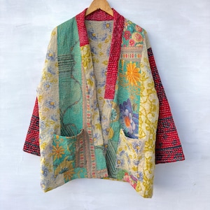 Könnte beinhalten: Eine langärmelige Jacke aus gestepptem Stoff mit einem Patchwork aus verschiedenen Blumen- und geometrischen Mustern in Rot-, Türkis-, Gelb- und Blautönen. Das Kleidungsstück verfügt über einen einfarbigen roten Kragen, zwei Vordertaschen und sichtbare Nähte.
