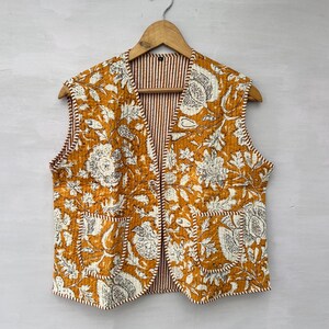 May include: Mustard yellow quilted vest with a floral pattern in white and gray. The vest has two front pockets and a striped red and white lining. It is hanging on a wooden hanger.