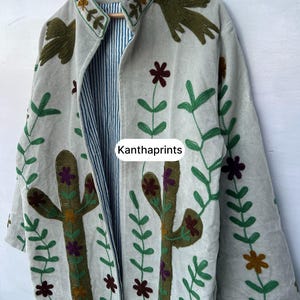 May include: A light-colored, open-front jacket with embroidered floral and cactus designs. The jacket features a striped lining and the text "Kanthaprints". The embroidery includes green, brown, and purple accents.