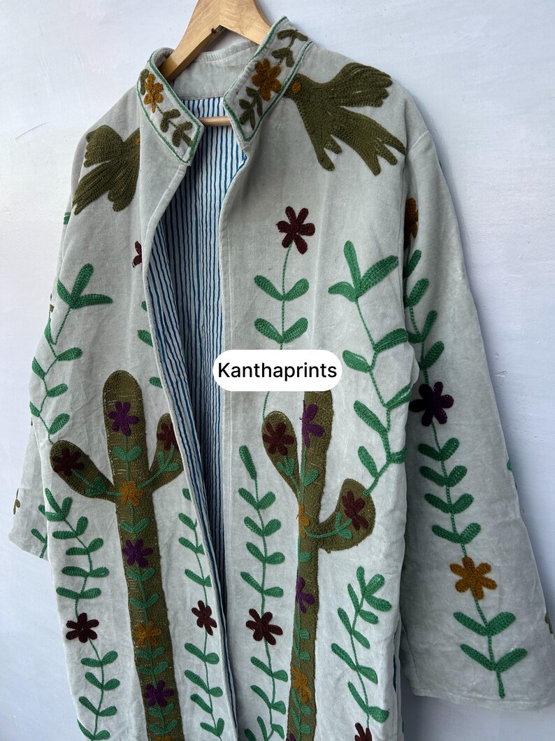 May include: A light-colored, open-front jacket with embroidered floral and cactus designs. The jacket features a striped lining and the text "Kanthaprints". The embroidery includes green, brown, and purple accents.