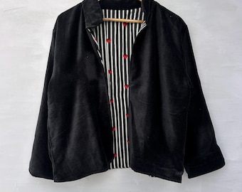 May include: A black velvet jacket hanging on a wooden hanger. The jacket has a collared neckline and long sleeves. The interior lining features a black and white striped pattern with small red hearts.