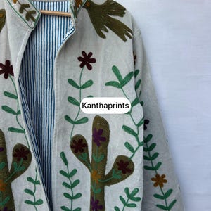 May include: Off-white jacket with embroidered floral and cactus designs in green, brown, and purple. The jacket has a striped blue and white lining and the text "Kanthaprints".