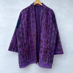 Puede incluir: Una chaqueta acolchada de color morado con un estampado oscuro y líneas de costura verticales en toda la prenda. Tiene mangas largas, un diseño frontal abierto y un pequeño detalle de parche azul oscuro en la manga derecha superior, colgada en percha.