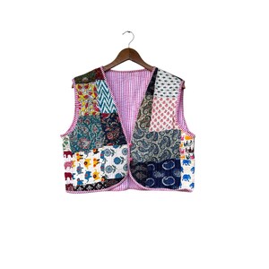 May include: A patchwork vest with a variety of colorful fabric patterns, including paisley, floral, and geometric designs. The vest has a pink striped lining and is hanging on a wooden hanger. The vest is sleeveless and has a V-neck.