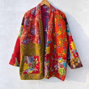 Può includere: Una giacca a maniche lunghe con un design patchwork caratterizzato da vivaci motivi floreali e geometrici nelle tonalità del rosso, giallo, verde e viola. Il capo presenta una tasca frontale, un collo a scialle e visibili impunture bianche.