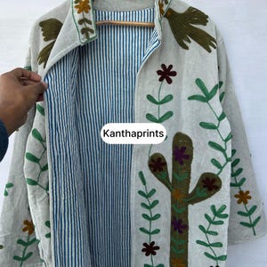 May include: Beige embroidered jacket with a blue and white striped interior. The jacket features floral and cactus embroidery in green, brown, and purple. The text "Kanthaprints" is visible on the front.